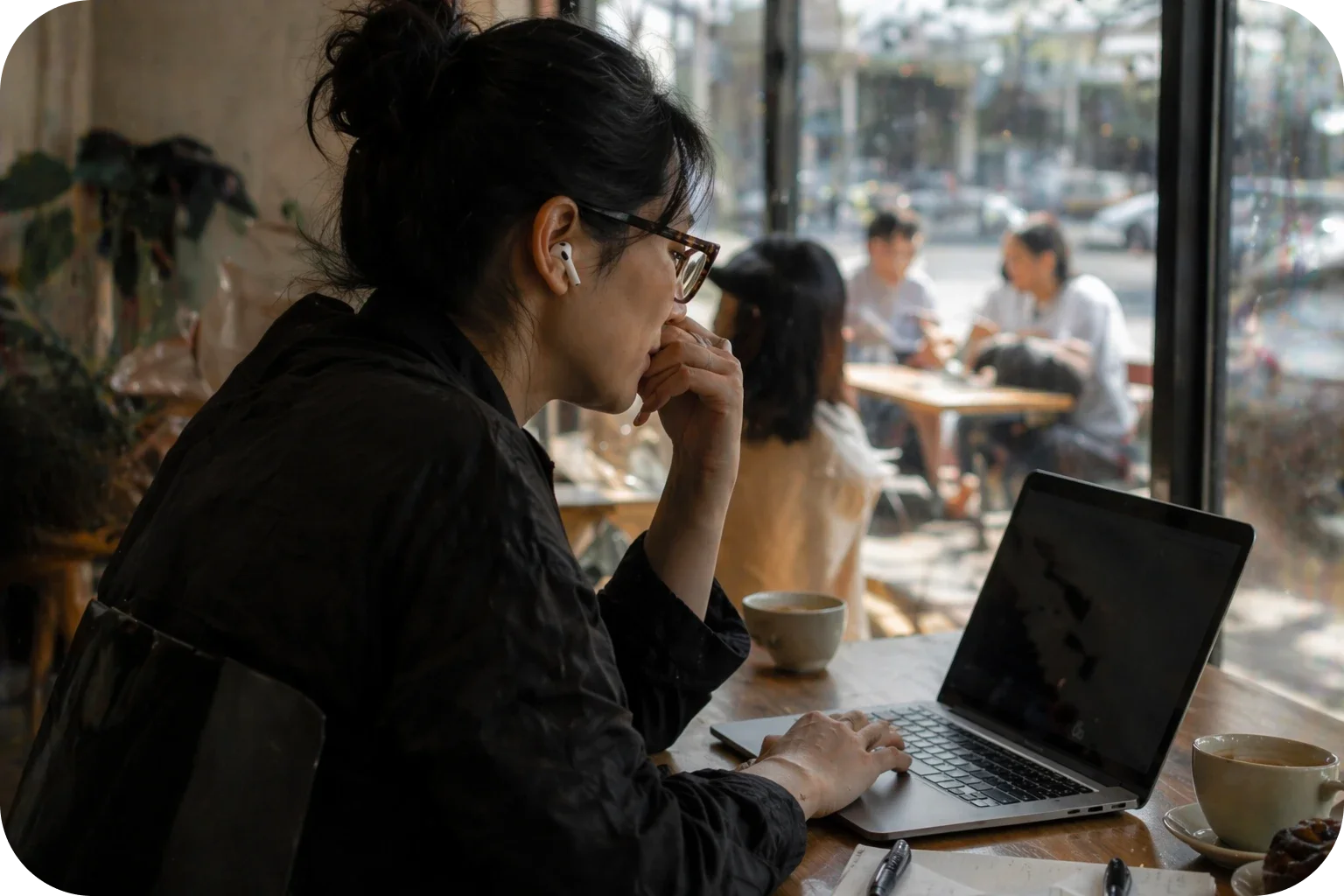 A person wearing earbuds uses a laptop beside a café window